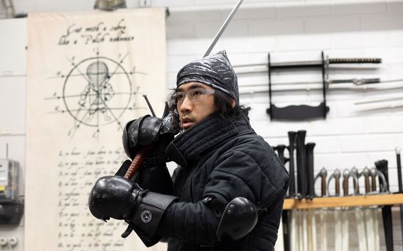 Russel Lai on kuvassa puettuna mustaa miekkailuasuun, pitää kädessään suurta miekkaa ja katsoo etuviistoon ylös kuin kohti vastustajaa. Taustalla näkyy miekkoja telineissä. 