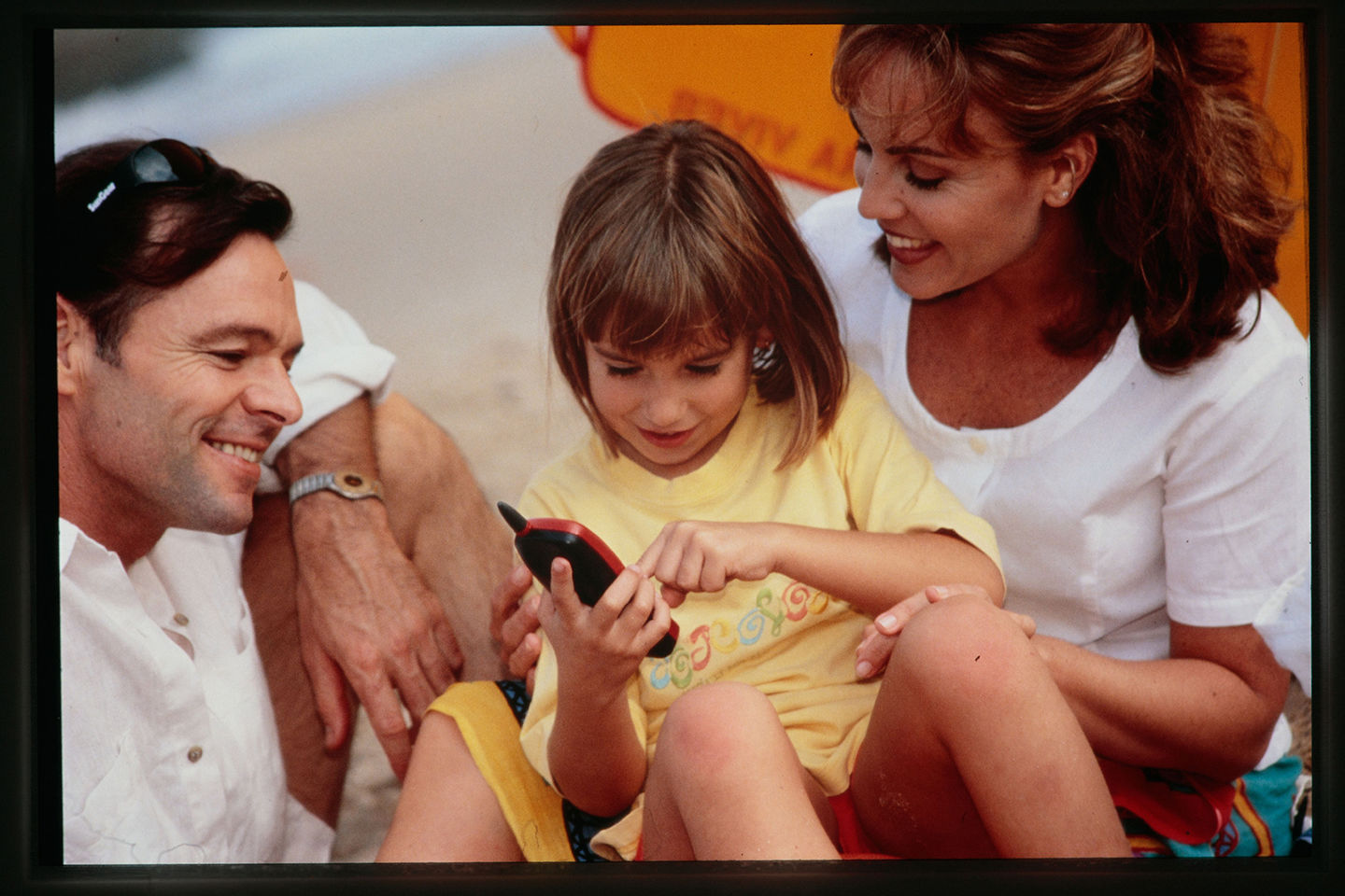 A child in a yellow shirt is using a mobile phone with two adults sitting beside them on a beach.