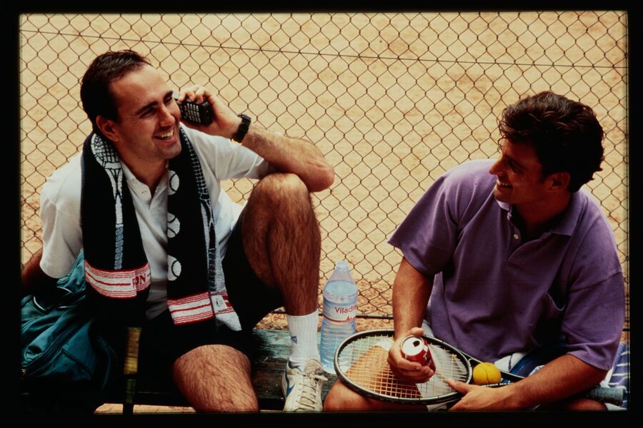 Two men sitting by a tennis court, one on the phone, the other holding a racket, with a water bottle and towel.