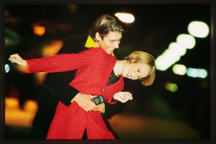 Two people, one in a red outfit, holding a phone, are posing closely together on a brightly lit street at night.