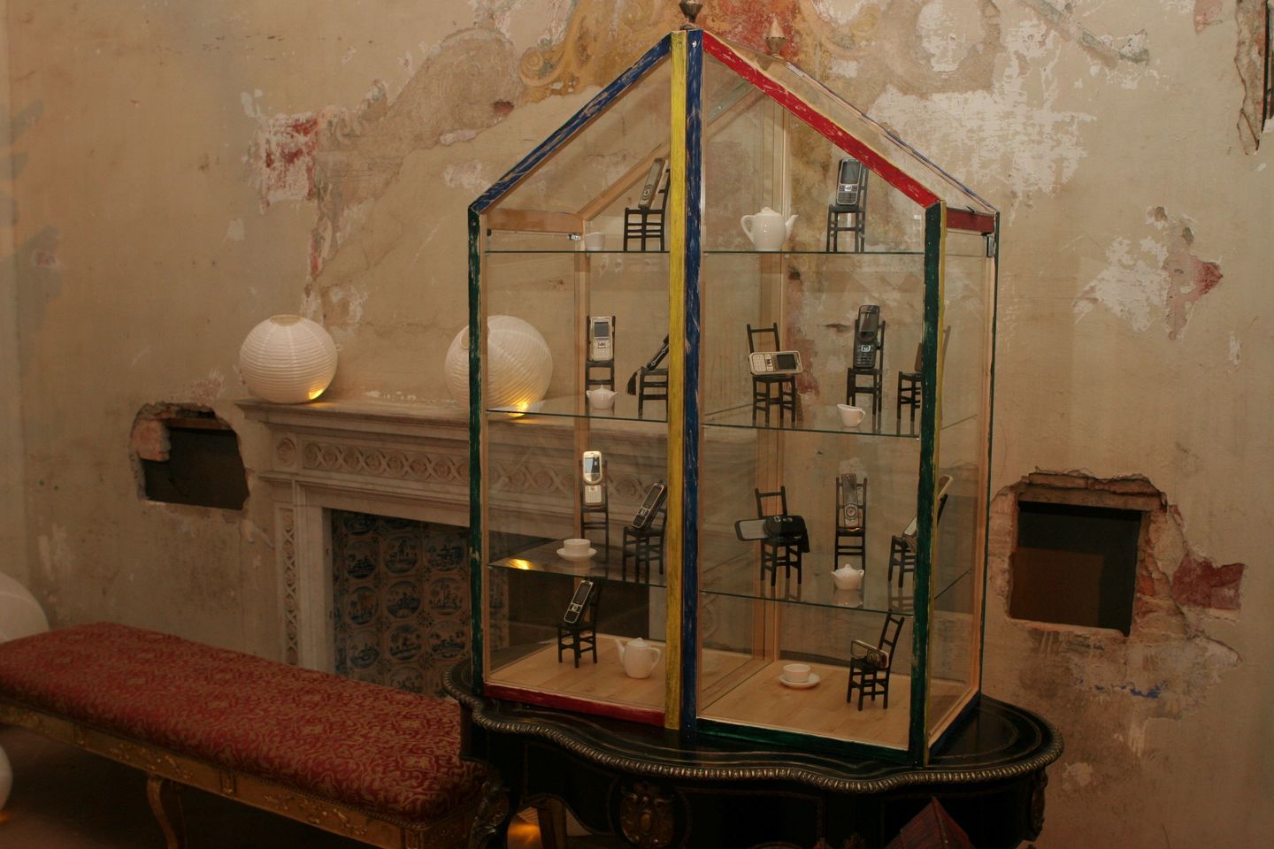 Glass display case shaped like a house with mobile phones on small chairs. Background shows an old wall and a fireplace.