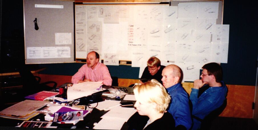 A group of people in a meeting room with technical drawings on the walls. Papers and materials are spread on the table.